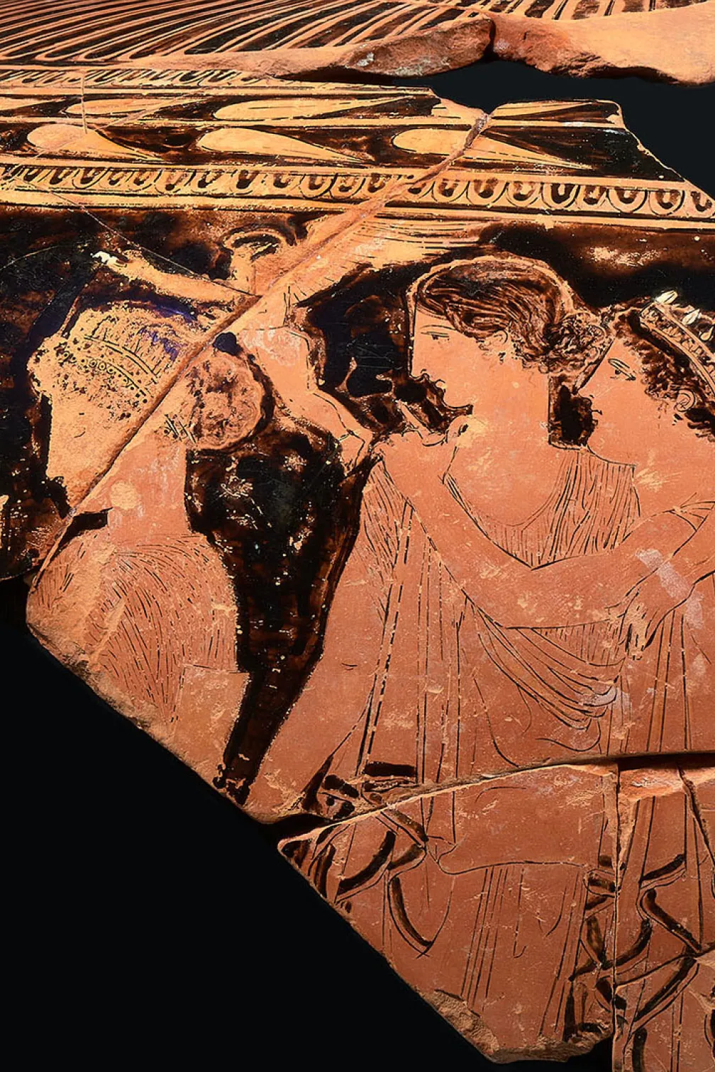 Ancient Greek terracotta pottery fragment showing two figures in profile with decorative patterns above.