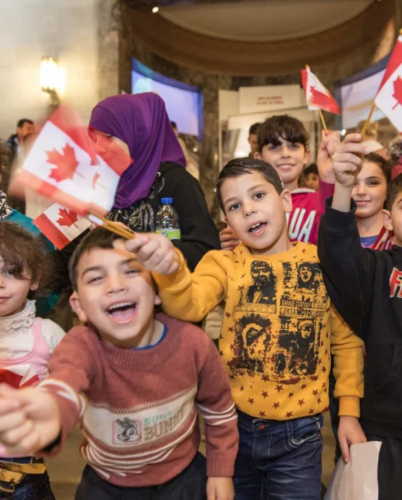 A group of Syrian children smile and wave Canadian flags in front of the ROMâs Schad Gallery of Biodiversity
