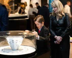 A young boy dressed in a suit is interacting with an exhibit under a glass dome while a woman in black watches.