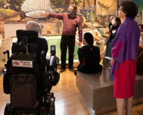 Museum guide wearing a face mask gesturing to visitors, including a person in a wheelchair, seated and standing, in a gallery with painted wall art.
