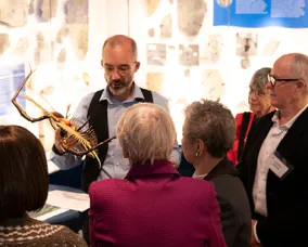 A man with glasses holds a large, antler-like object while explaining it to a small group of older adults.