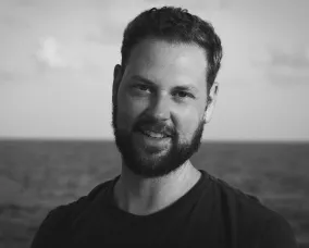 Un homme caucasien souriant, aux cheveux et à la barbe foncés, vêtu d'un t-shirt noir.