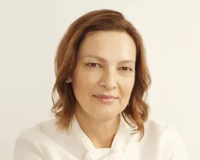 Portrait of a woman with light brown hair, wearing a white blouse, against a plain beige background.