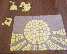Un soleil en carton est posé sur une table. Le centre du soleil et la moitié de l'espace entre les rayons du soleil sont remplis de carrés jaunes. D'autres piles de carrés jaunes se trouvent sur la table à côté du dessin.