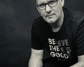 Man with short gray hair and glasses wearing a black shirt with the white text "BELIEVE THERE IS GOOD."