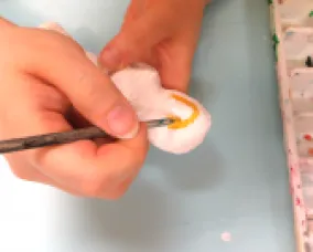 Close-up of hands painting a small pink object using a thin paintbrush near a watercolor palette.