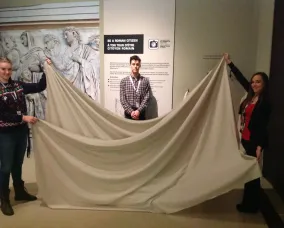 Two young women hold up a long piece of white fabric in front of a young man.