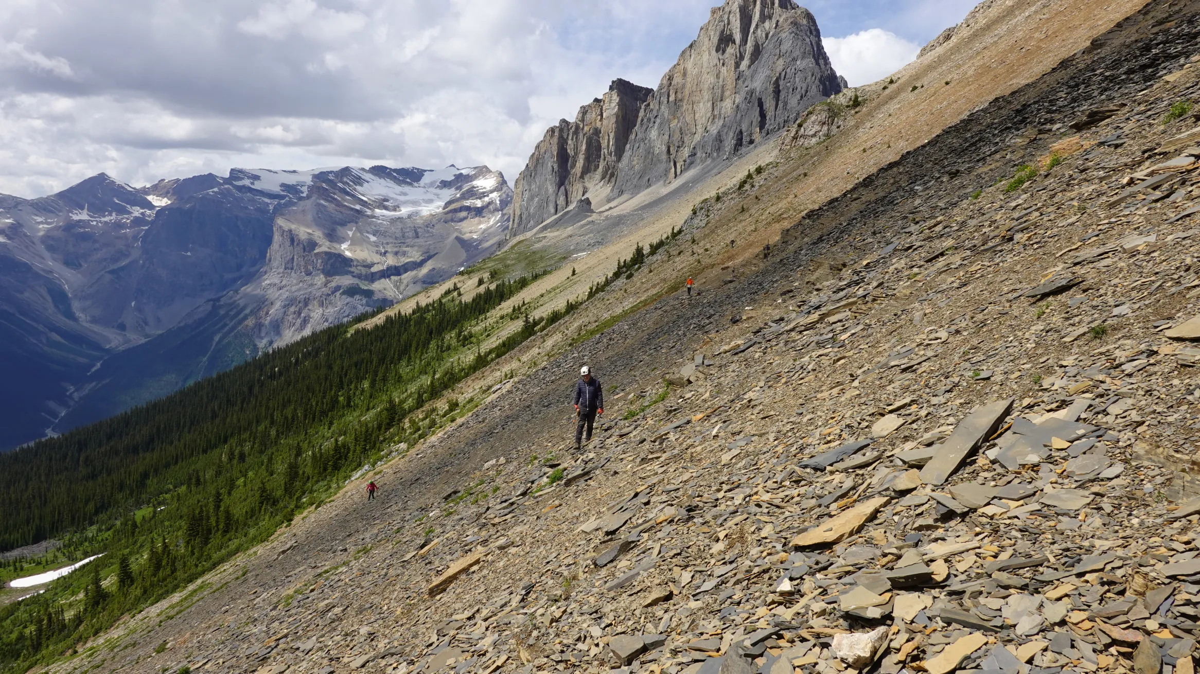 Hiker walking on a steep rocky mountain slope with rugged peaks and cloudy sky in the background.