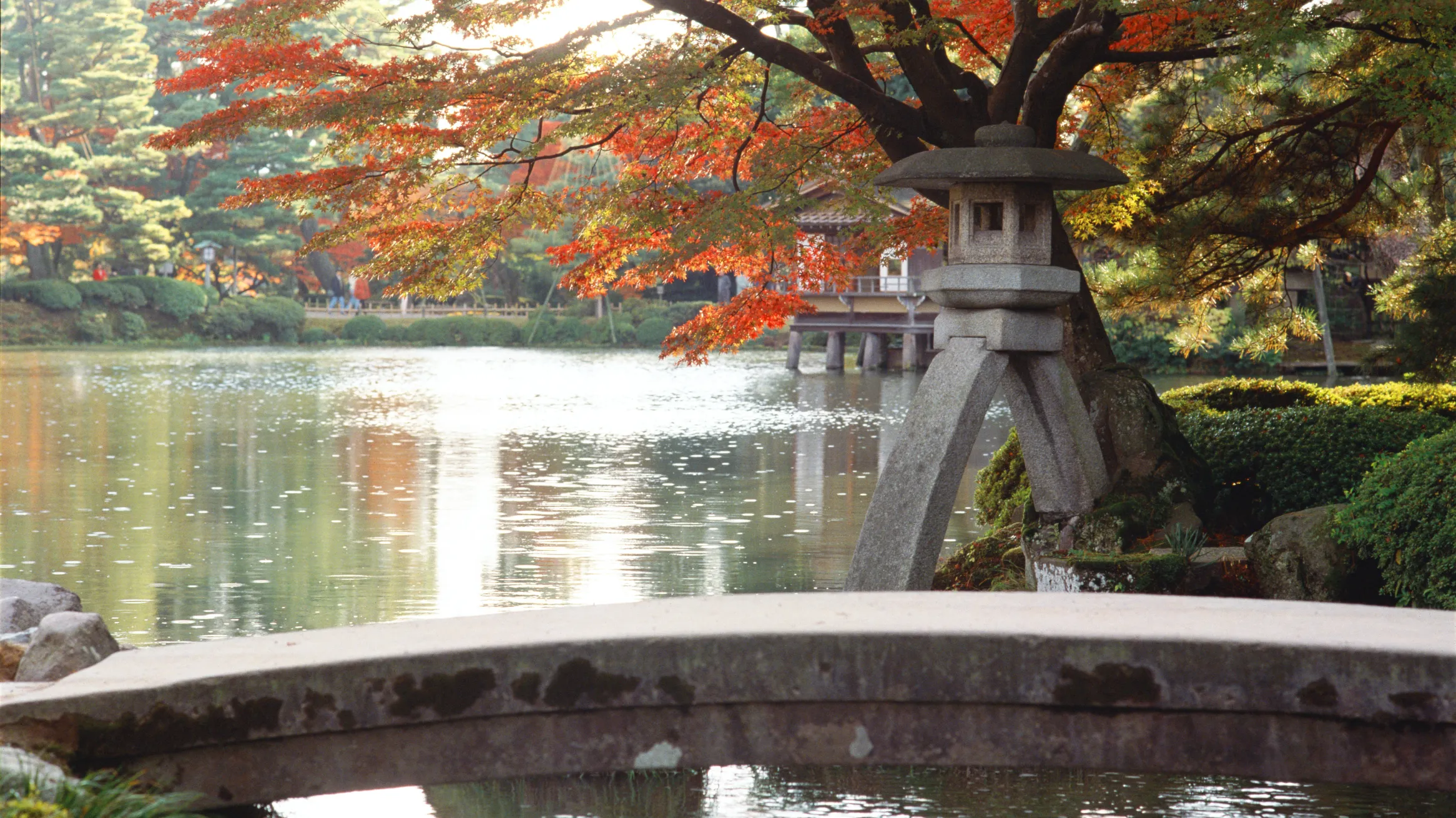 Stone bridge over a pond with autumn-colored trees and a traditional Japanese stone lantern beside the water.