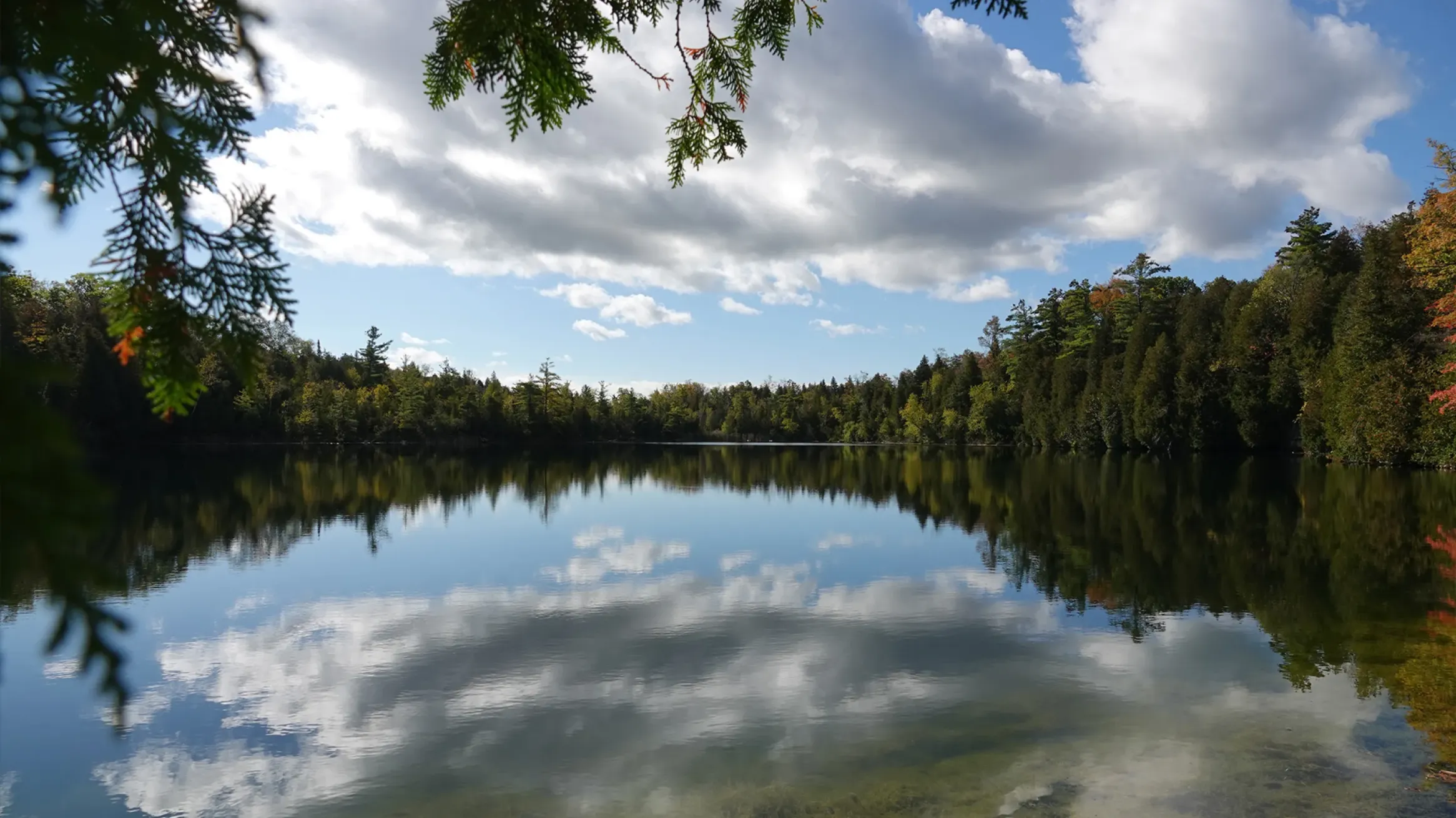 Calm lake reflecting a sky with white clouds, surrounded by dense forest trees on a partly sunny day.